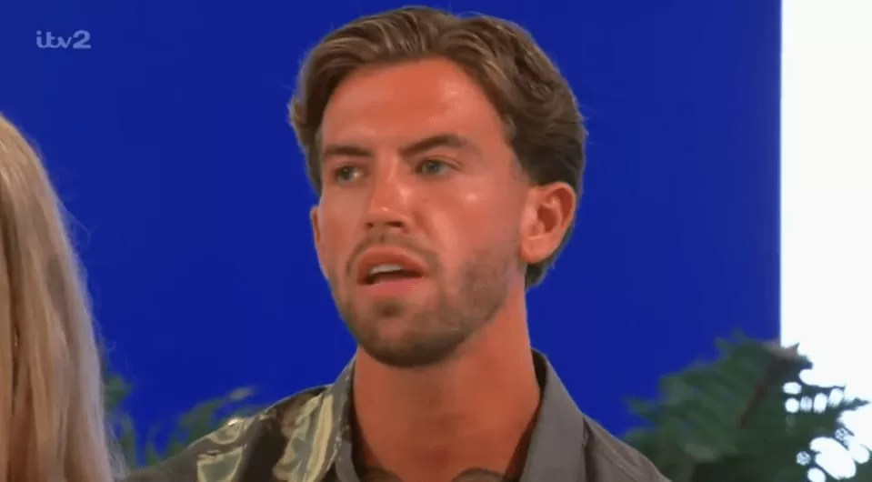 A man with styled brown hair and a beard, wearing a patterned shirt, looking distressed with his mouth slightly open.