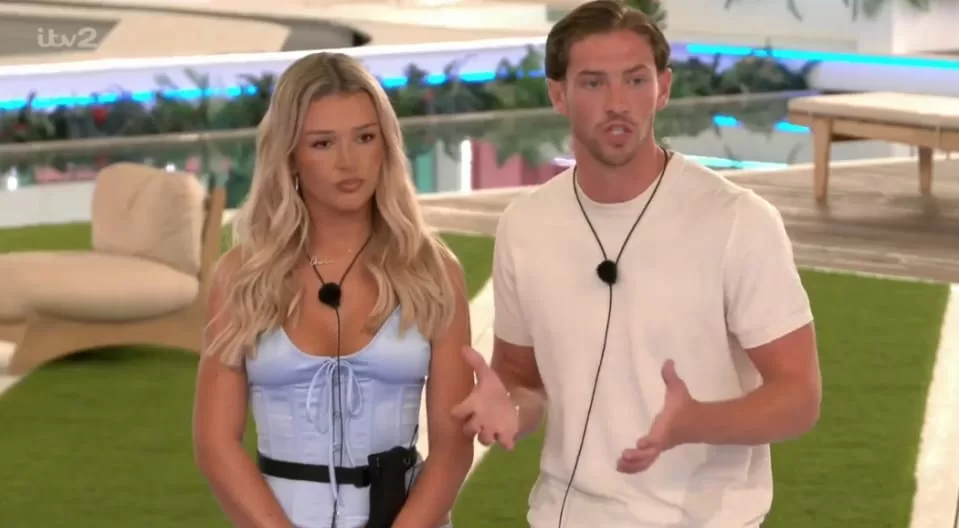 A man with a white t-shirt and a woman with a light blue corset top, both wearing microphones, stand outdoors on what appears to be a reality television set, speaking to someone off-camera.